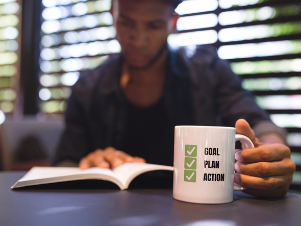 Man reading a book holding a coffee cup that reads GOAL PLAN ACTION